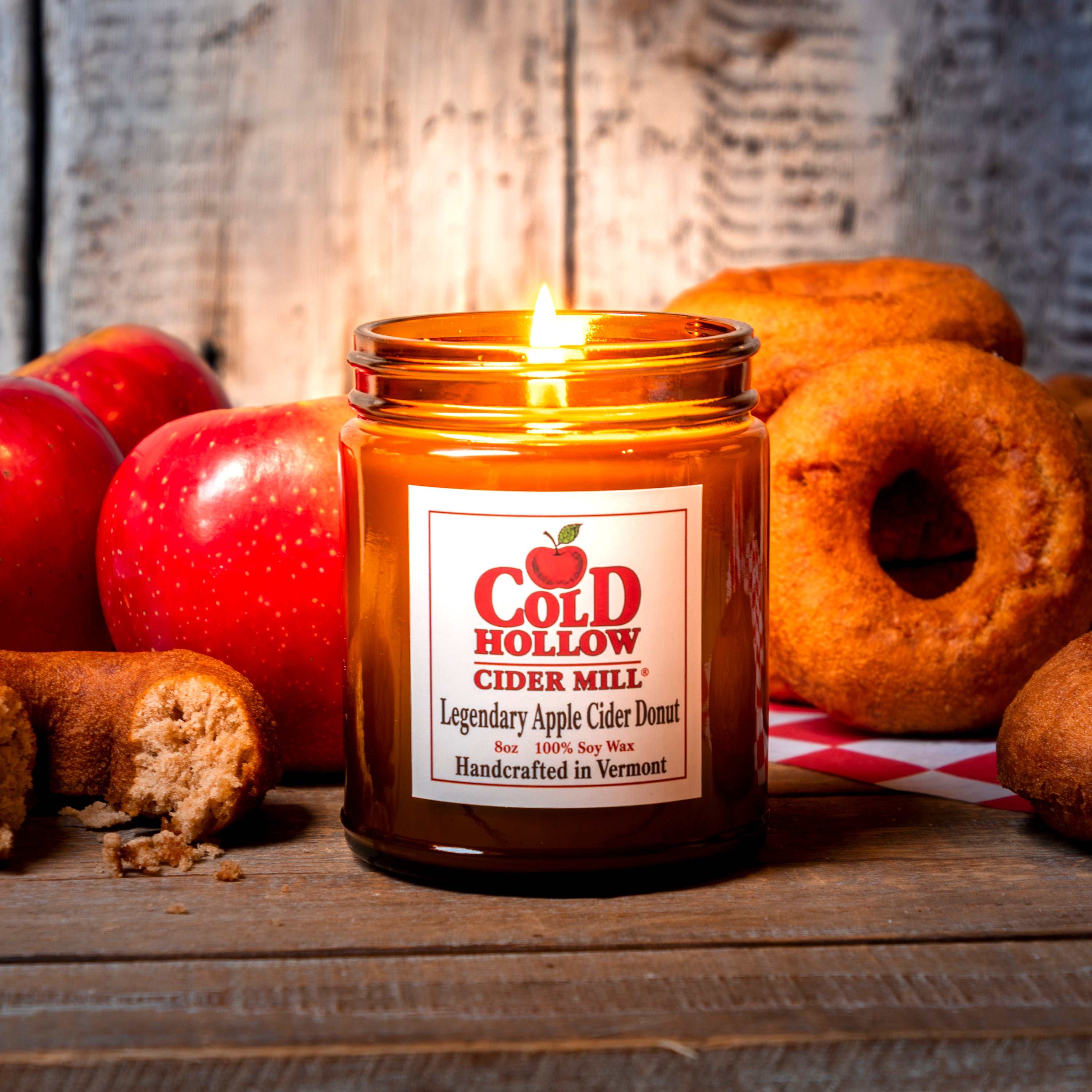 Candle in a jar labeled 'Cold Hollow Cider Mill' with apples and donuts on a wooden surface.