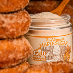 A jar of Cold Hollow Pure Vermont Maple Sugar surrounded by sugar-coated doughnuts, with a wooden spoon resting in the open jar.