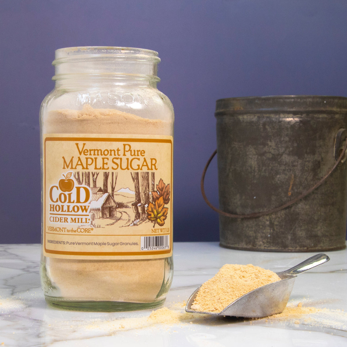 A glass jar of Vermont Farm Maple Sugar labeled 'Cold Hollow Sugar White' stands on a white marble surface with a scoop of the sugar in front and a dark metal bucket behind, against a blue backdrop.