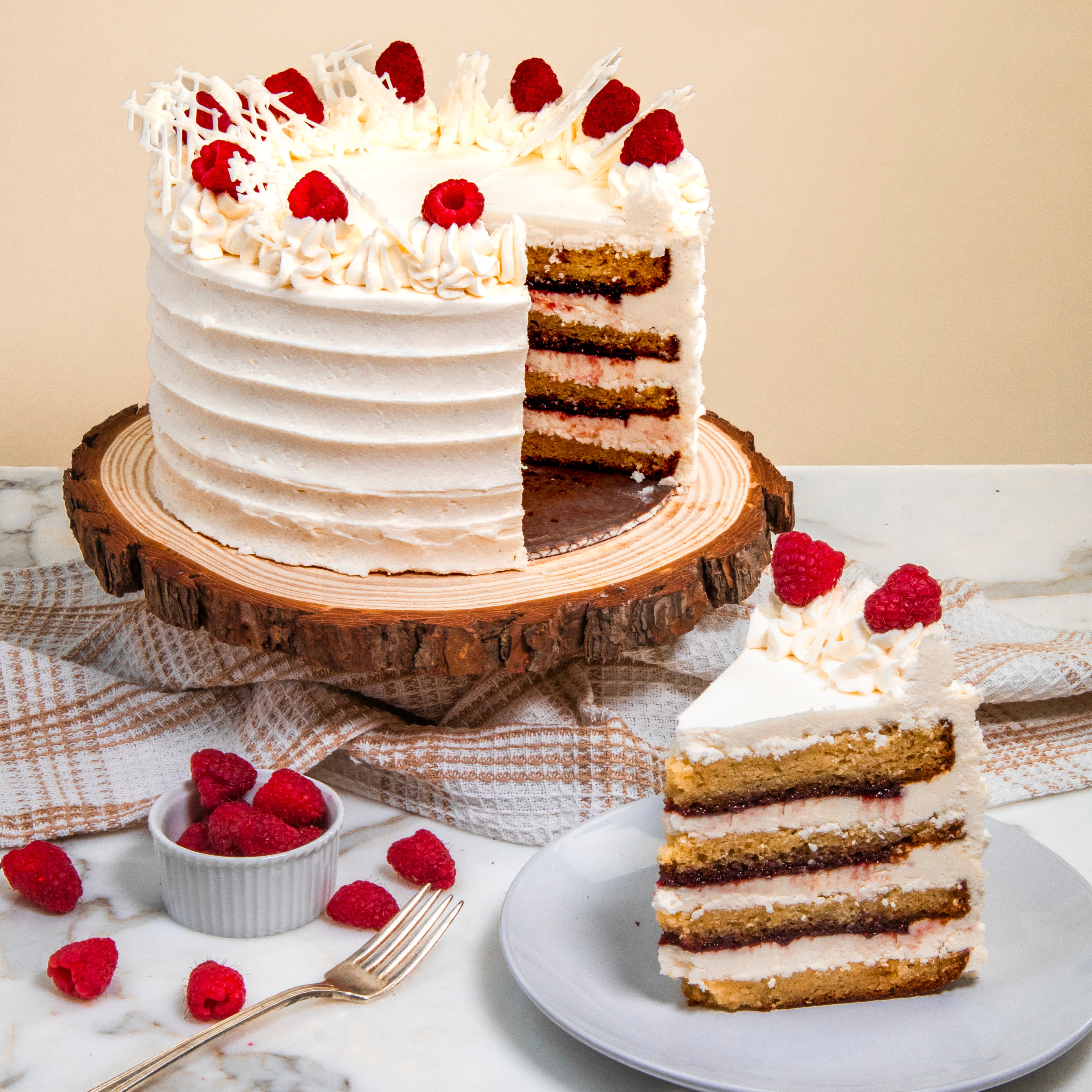 Layered cake with white frosting and raspberries on a wooden cake stand, with a slice on a plate.