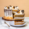 A layered cake with white frosting and chocolate drizzle sits on a wooden slice, with one slice cut and placed on a plate in front. The cake is topped with whipped cream, chocolate pieces, graham cracker crumbs, and toasted marshmallows, and set on a marble surface against a beige background.