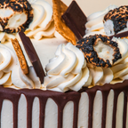 Close-up of a frosted cake with white piped swirls, dark chocolate drizzle, graham cracker pieces, and toasted marshmallow treats on top.