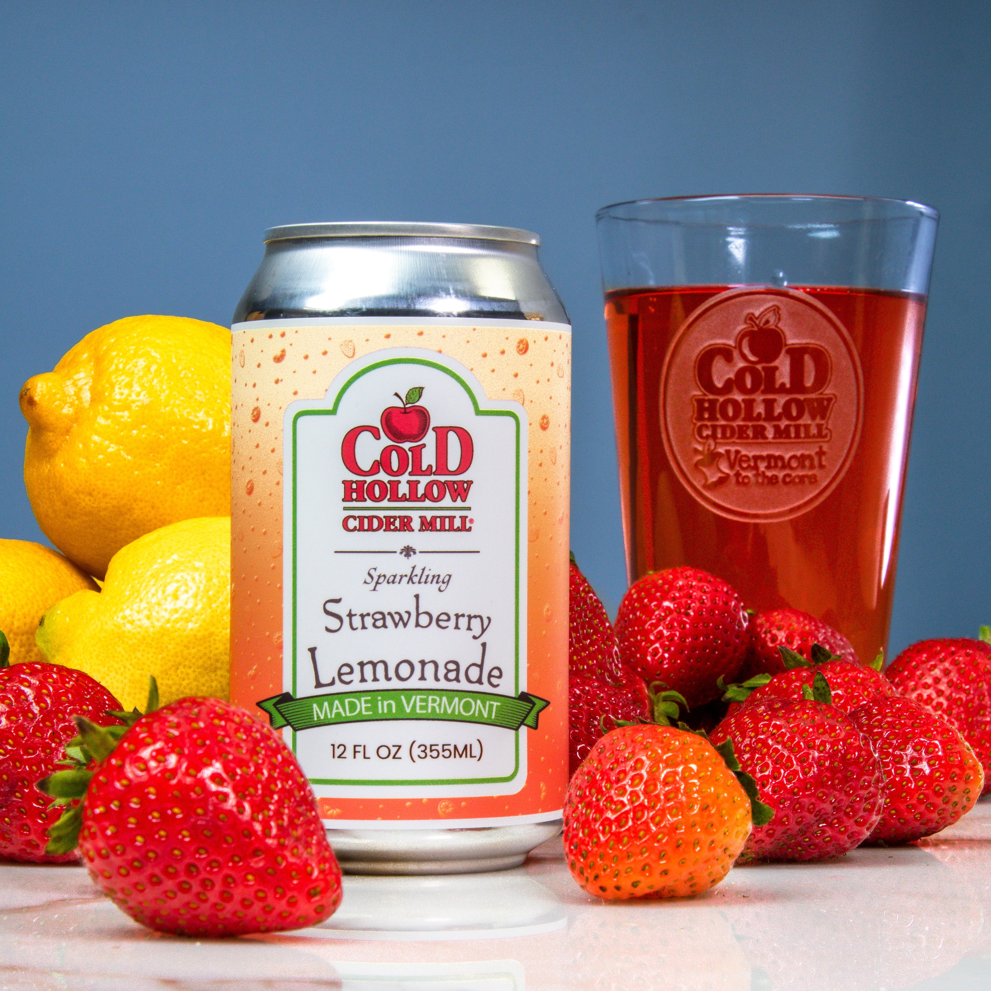 Can of Cold Hollow Cider Mill Strawberry Lemonade with a glass of cider, lemons, and strawberries on a reflective surface.